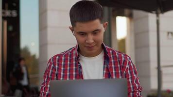 frente Visão do uma jovem ásia homem fechar acima sentado às a ao ar livre cafeteria e trabalhando com entusiasmo em uma computador portátil com uma construção dentro uma pano de fundo. conceito do moderno controlo remoto trabalhos com uma flexível cronograma video