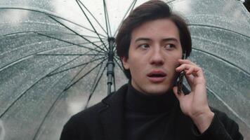 Front view of a young man in a coat walking under a transparent umbrella during a rain in the autumn city park and talking on a smartphone with a friend video