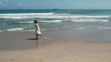 jovem adulto mulher dentro uma branco vestir alegremente corrida ao longo uma arenoso de praia litoral, com □ Gentil ondas e distante barcos em uma ensolarado dia, transmitindo liberdade e terra dias conexão para natureza video