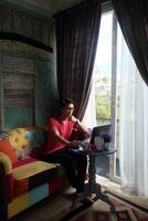 Asian Man Working on Laptop in Sunlit Villa, Sofa Comfort, Productivity photo