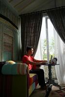 asiático hombre trabajando en ordenador portátil en iluminado por el sol villa, sofá comodidad, productividad foto