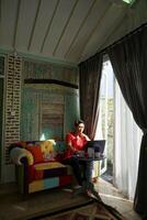 Asian Man Working on Laptop in Sunlit Villa, Sofa Comfort, Productivity photo