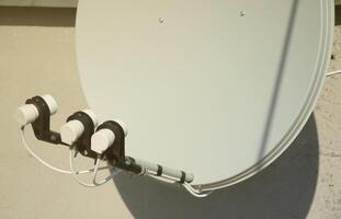 White satellite dish with three converters mounted on residental building rooftop concrete wall. Satellite television photo