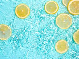 Lemon slices in clean transparent water over blue background with copy space. Water splashing on blue water surface in sunlight. Top view or flatlay. Summer, vacation, healthy eating concept photo