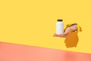 Empty white medical container for vitamins in female hand with sprinkle manicure through torn bright yellow paper background. Oral supplements, immune support, biohacking creative concept. Copy space photo