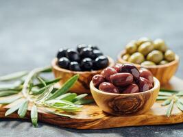 Set of green, red and black olives on gray background. Different types of olives in olive wooden bowls and olive oil over wooden cutting board and fresh olive leaves. Copy space. photo