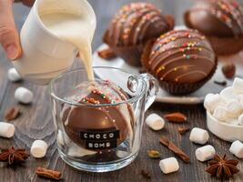 Chocolate cocoa bomb in glass cup with choco bomb text. Pouring plant-based hot milk on ball made from milk chocolate with marshmallow inside. Trendy winter hot chocolate drink. Action shot photo