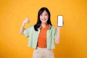 young Asian woman in her 30s, wearing orange shirt and green jumper, shows smartphone screen display with a thumbs-up gesture on yellow studio background. New mobile application concept. photo