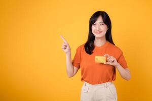 Energetic asian woman in orange shirt pointing finger to a credit card, highlighting efficient online payment services. photo