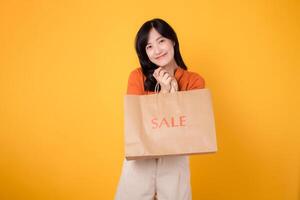 Express the thrill of savings and shopping with an attractive asian woman holding a bag isolated on yellow background. Youthful and trendy retail concept. photo