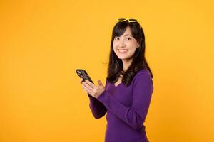 alegre joven asiático mujer 30s vistiendo púrpura camisa contento cara utilizando un móvil teléfono aislado en amarillo antecedentes. móvil teléfono tecnología concepto. foto