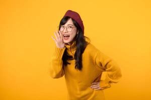 Happy young Asian woman wearing yellow sweater with open mouths raising hands screaming announcement isolated on yellow background photo