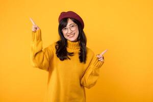 joven asiático mujer en su 30, vistiendo un amarillo camisa y rojo boina. contento cara y señalando dedo a gratis Copiar espacio en contra vibrante amarillo fondo. desbloquear ahorros y tienda ahora foto