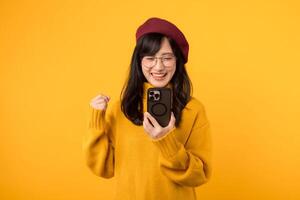 Happy joyful young 30s asian woman wearing yellow shirt standing using mobile cell phone typing sms message doing winner gesture isolated on yellow colour background, studio portrait photo