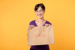 Pretty cheerful attractive Asian 30s young woman wearing purple shirt with happiness face holding sale shopping paper bag isolated on yellow background. Shopaholic woman concept. photo