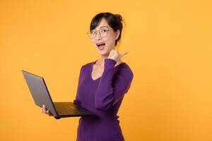 retrato alegre joven 30s asiático mujer contento sonrisa vistiendo púrpura camisa y lentes utilizando ordenador portátil computadora mientras señalando dedo a gratis Copiar espacio aislado en amarillo antecedentes. foto