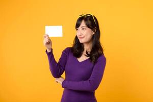 Portrait young pretty Asian woman 30s wearing purple shirt sweater while showing blank empty paper board card in her hand isolated on yellow background. Business presentation post card message concept photo