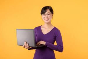 Portrait pretty young 30s Asian female student or businesswoman wearing purple sweater with glasses using a laptop computer for online meeting work isolated on yellow background. meeting concept. photo