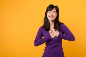 Portrait pretty young Asian 30s woman wearing purple shirt white holding hands on chest isolated on yellow background. Thankful, healthy wellness life concept. photo