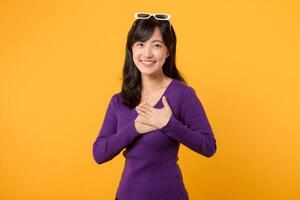 Portrait pretty young Asian 30s woman wearing purple shirt white holding hands on chest isolated on yellow background. Thankful, healthy wellness life concept. photo