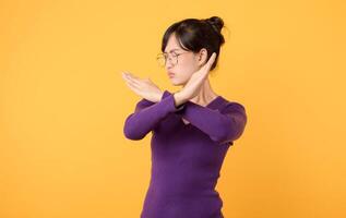 serious lady wearing purple shirt and eyeglasses, using X sign arms to convey message of rejection or prohibition against a yellow background. Explore the power of non-verbal communication. photo