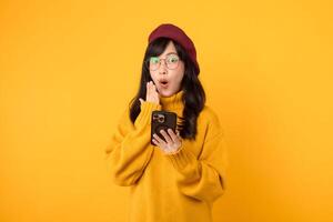 Surprised young asian 30s woman using mobile phone with positive expression, smiles broadly, dressed in yellow shirt and standing isolated on yellow background. Happy adorable glad rejoices success. photo