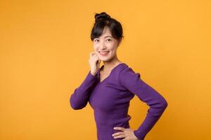 Portrait young cheerful asian woman 30s wearing purple shirt happy smile while holding her hip isolated on yellow background. photo