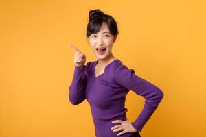 Portrait cheerful young asian woman 30s wearing purple shirt pointing finger to free copy space isolated on yellow background. photo