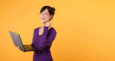 Asian female teacher 30s cute smile holding laptop pointing finger to free copy space, Wearing purple sweater shirt and standing in a yellow studio setting. Exchange education advertising concept. photo