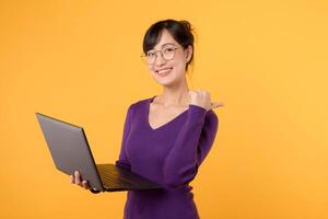 Portrait young asian woman student wearing purple shirt and eyeglasses happy smile holding laptop while pointing thumb to free copy space against yellow background. education concept. photo