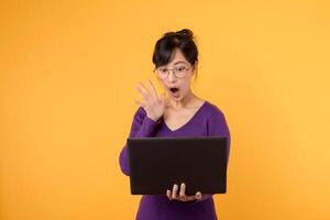 Young surprised woman of Asian ethnicity 30s wear purple shirt with glasses  using laptop pc computer isolated on plain yellow studio background. Surprise news from online electronic mail concept. photo