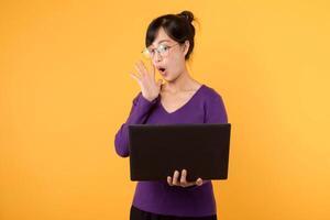 Portrait young asian woman student wearing purple shirt and eyeglasses surprise face holding laptop isolated on yellow background. education, businesswoman model concept. photo