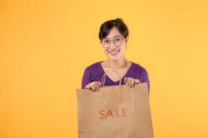 Pretty cheerful attractive Asian 30s young woman wearing purple shirt with happiness face holding sale shopping paper bag isolated on yellow background. Shopaholic woman concept. photo