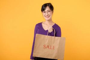 A confident and cheerful female shopper, donning a stylish purple shirt and holding a shopping bag against a yellow background, signifies a great shopping experience. photo