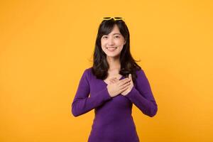 Against yellow background, a young woman, wearing a purple shirt and eyeglasses, sends cheerful and loving Valentine's Day gesture with her hand, expressing happiness and romance in a studio portrait. photo