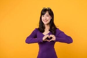 Against a yellow background, expressing love and happiness, a cheerful young woman, wearing a purple shirt and eyeglasses, forms a heart shape with her hand in a studio portrait, symbolizing romance. photo