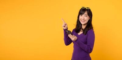 Portrait young asian pretty woman 30s happy smile wearing purple shirt and sunglasses pointing finger to free copy space and holding smartphone against yellow background. new app mobile concept photo