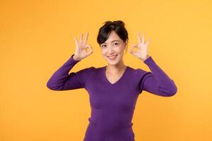 Okay sign gesture. Young 30s asian woman wearing purple shirt doing OK hand gesture with happy confident smile isolated on yellow background. Agree, Okay, approved hand gesture concept. photo