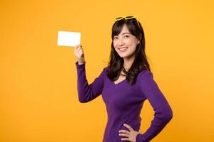 Beautiful pretty smiling asian woman 30s wearing purple shirt showing blank gift card isolated on yellow background. photo