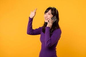 Vibrant Asian woman in her 30s, wearing purple shirt and sunglasses, using smartphone with okay hand sign on vibrant yellow background. New mobile app concept. photo