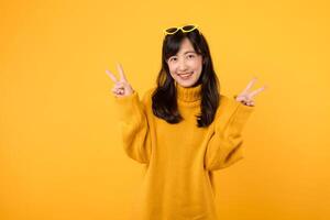 Portrait young asian woman 30s happy smile wearing yellow sweater and yellow sunglasses showing two fingers victory hand sign gesture isolated on yellow background. photo