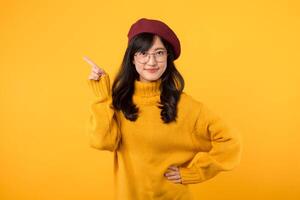 An elegant lady donning a yellow sweater, red beret, and eyeglasses, radiating positivity while pointing her finger to free copy space against a sunny yellow backdrop. photo