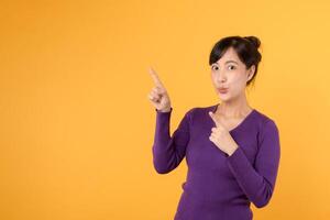 Look. Young pretty 30s asian woman wearing purple shirt expression pointing finger to free copy space for showing product isolated on yellow background. Advertisement concept. photo