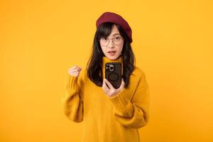 utilizando su teléfono inteligente con un puño en alto gesto. un alegre mujer, en un amarillo suéter y rojo boina, celebra en contra un vibrante amarillo antecedentes. foto