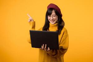 Attractive woman in corporate attire, confidently pointing to free copy space on her laptop, dressed in a yellow sweater and red beret against vibrant yellow background. photo