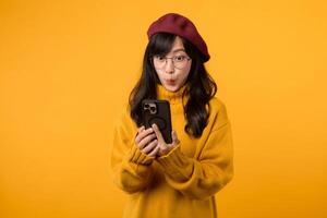 Portrait young Asian woman 30s wearing yellow shirt and glasses receive an exited news from mobile phone isolated on yellow background. Great news concept. photo
