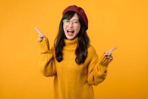 joven asiático mujer en su 30, vistiendo un amarillo camisa y rojo boina. contento cara y señalando dedo a gratis Copiar espacio en contra vibrante amarillo fondo. desbloquear ahorros y tienda ahora foto