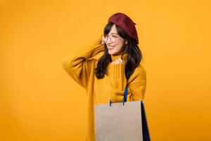 un contento joven mujer en un rojo boina y amarillo suéter disfruta su compras juerga, participación un papel bolso en contra un vibrante amarillo antecedentes. foto