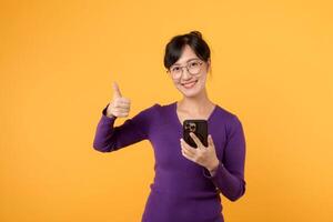 smiling young woman wearing purple shirt and eyeglasses, thumb up gesture while using her smartphone for online shopping and social media. Embrace joy digital communication against yellow background. photo