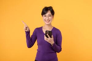 Portrait young asian pretty woman 30s happy smile wearing purple shirt and eyeglasses pointing finger to free copy space and holding smartphone against yellow background. new app mobile concept photo